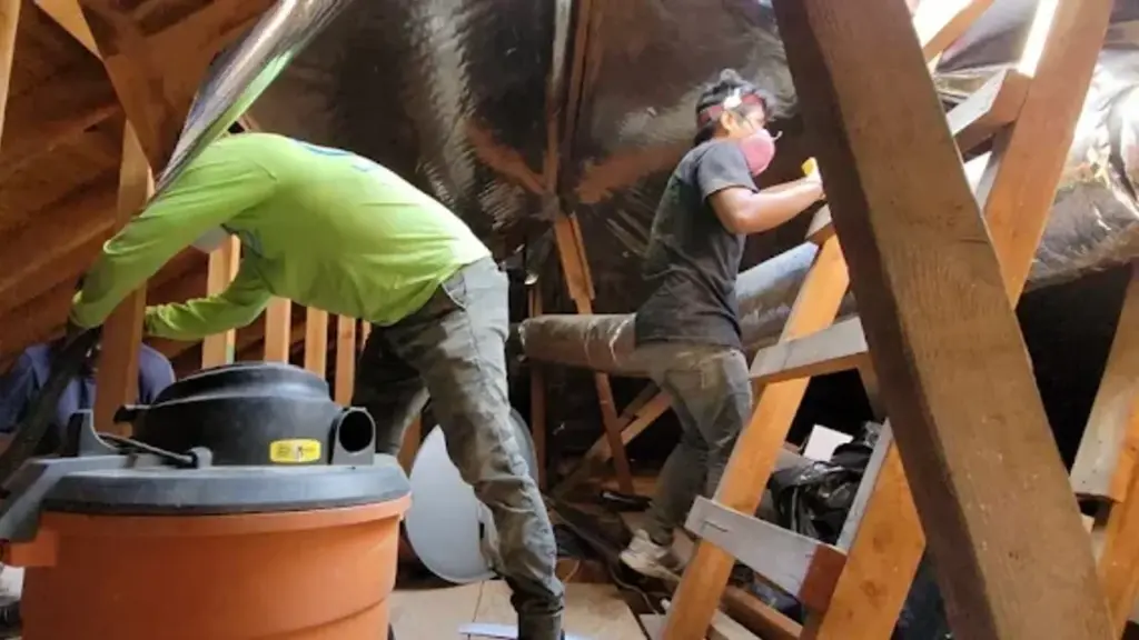 A wide-angle photograph taken inside a sweltering Los Angeles attic on a bright summer day, showing silver flexible duct
